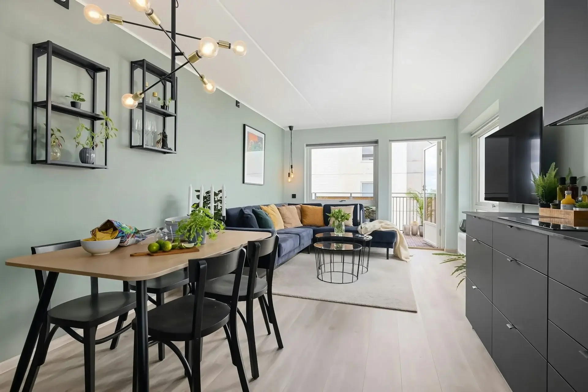Bright modern living and dining area with open-plan layout, wooden dining table, black chairs, cozy sofa, indoor plants, and balcony access.