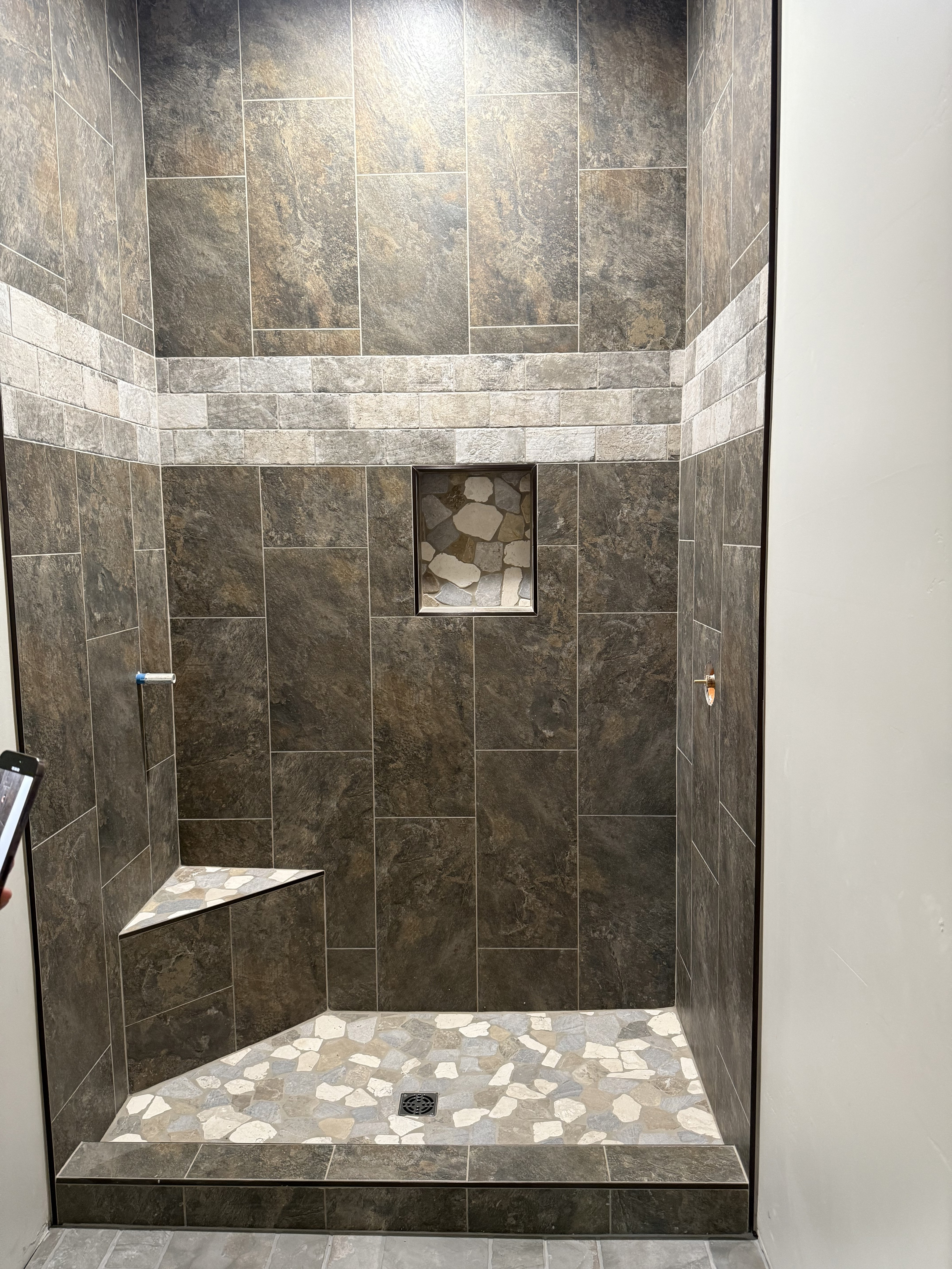 Spacious walk-in shower with dark grey rectangular wall tiles, a contrasting light pebble mosaic shower floor, and light grey floor tiles in the surrounding bathroom.