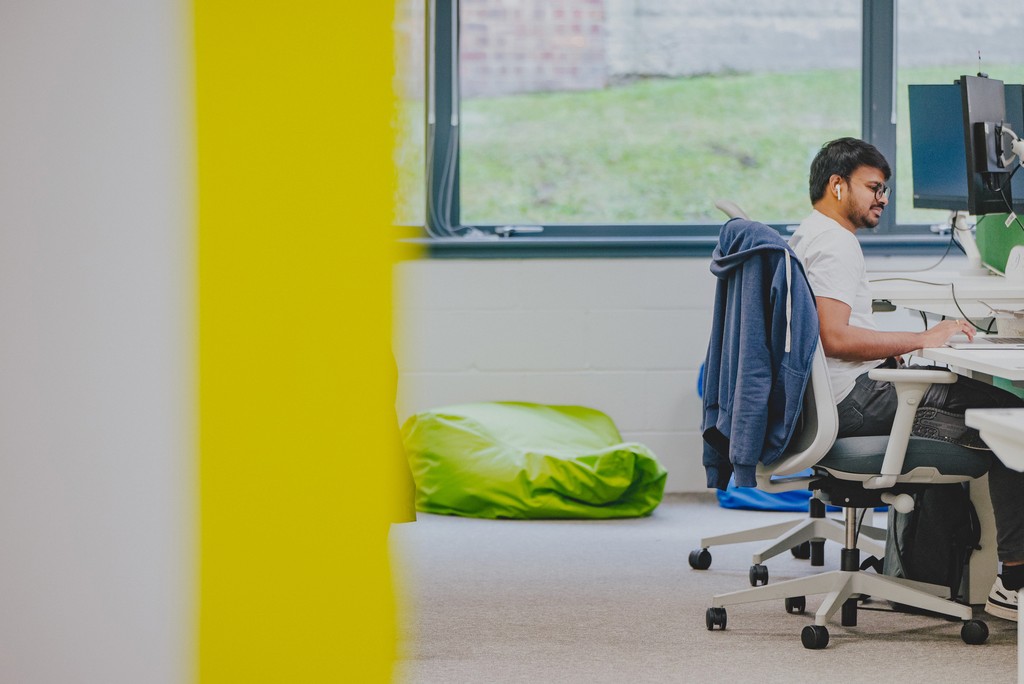 A modern office workspace with a person seated at a desk using a computer, positioned near a large window that lets in natural light. The scene includes rolling office chairs, a desk with monitors and equipment, a jacket draped over a chair, and a bright green beanbag on the carpeted floor. A bold yellow wall frames the left side of the image, adding contrast to the neutral interior.