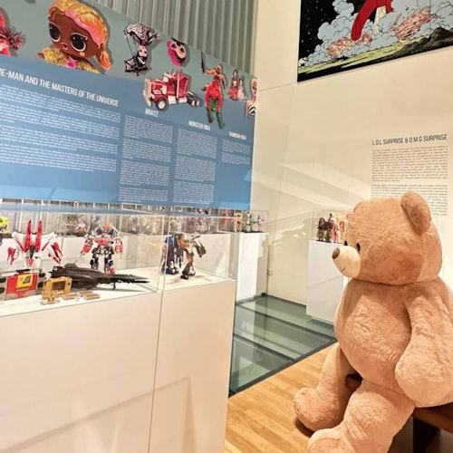 A museum exhibit displaying various action figures and toys, with an oversized teddy bear seated in the foreground.