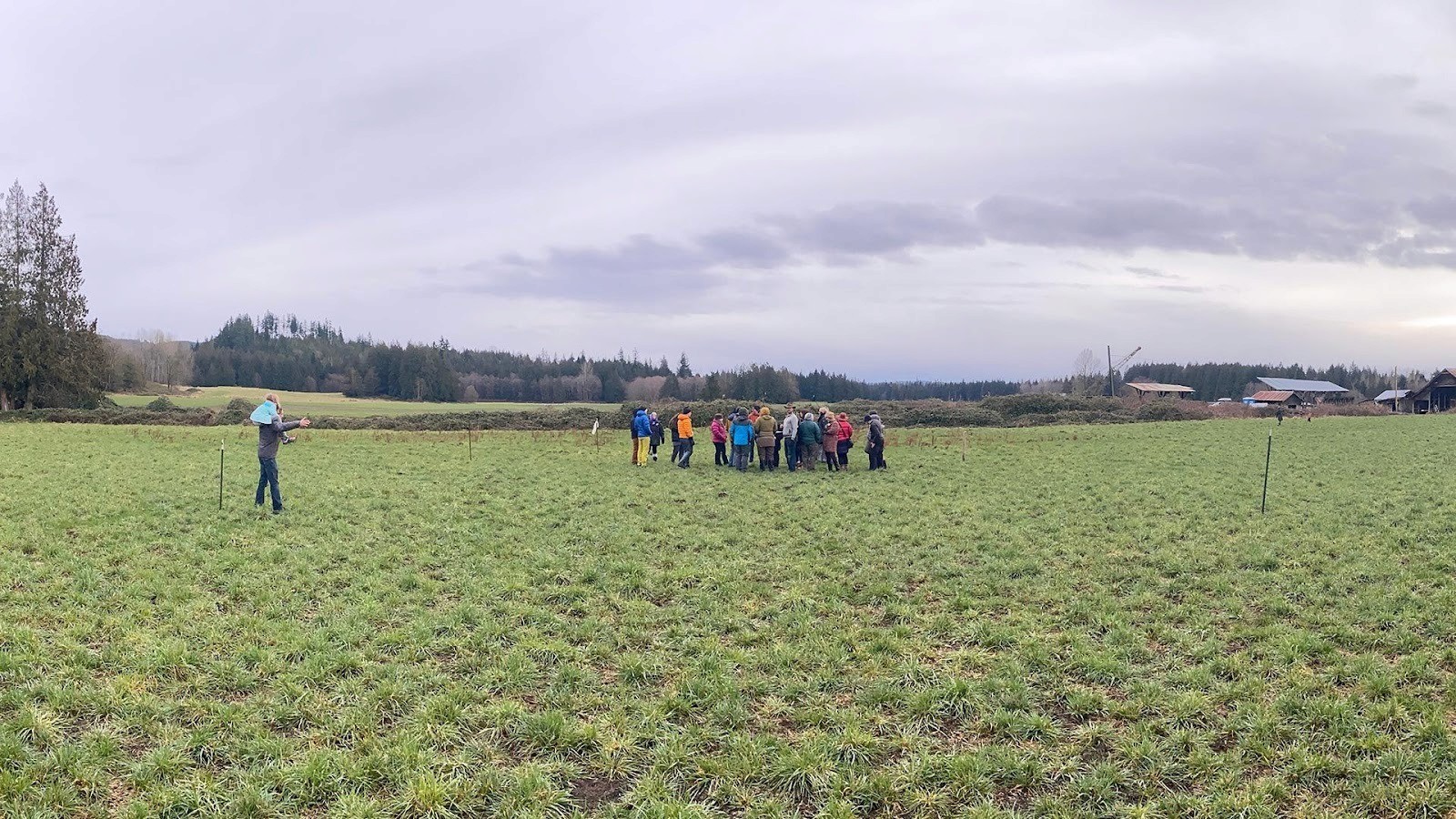 Group of future neighbors gathered together in an open grassy field at Rooted Northwest, with farmland and forest in the distance.