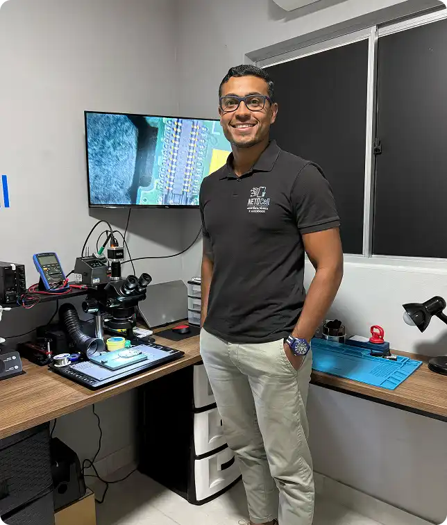 Profissional sorridente na sua assistencia técnica de celulares NETOCell, vestindo polo preta com logo, em bancada com microscópio de inspeção, multímetro e monitor mostrando placa eletrônica — reparo e assistência técnica de celulares.