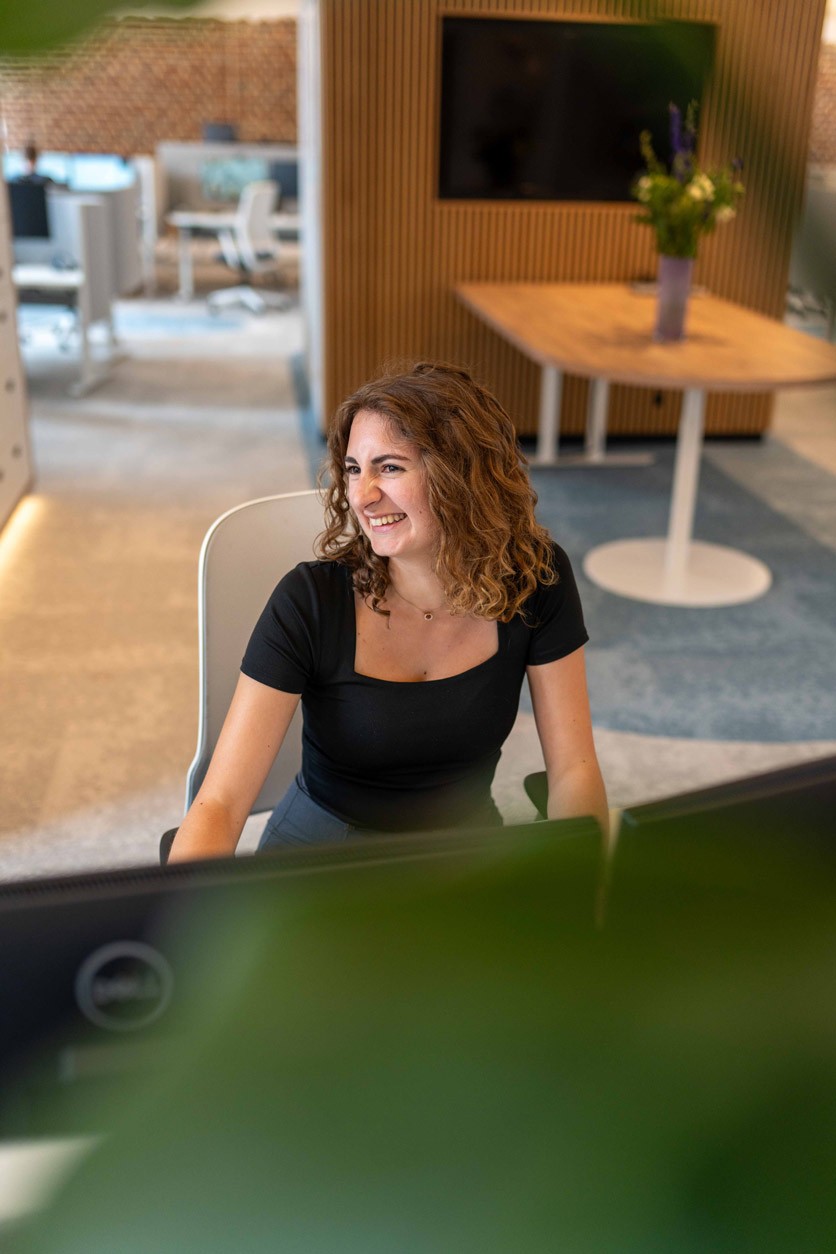 Mitarbeiterin mit lockigem Haar sitzt lächelnd am Schreibtisch in einem modernen Büro von SLB.