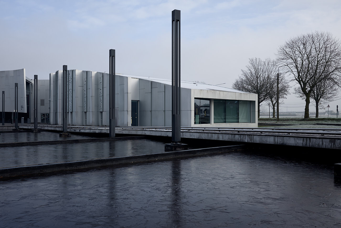 Photographie large d'architecture extérieure du Centre Culturel de Lesquin : façade moderne en béton blanc et rythmée se reflétant dans un bassin d'eau gelé sombre avec des luminaires verticaux graphiques.