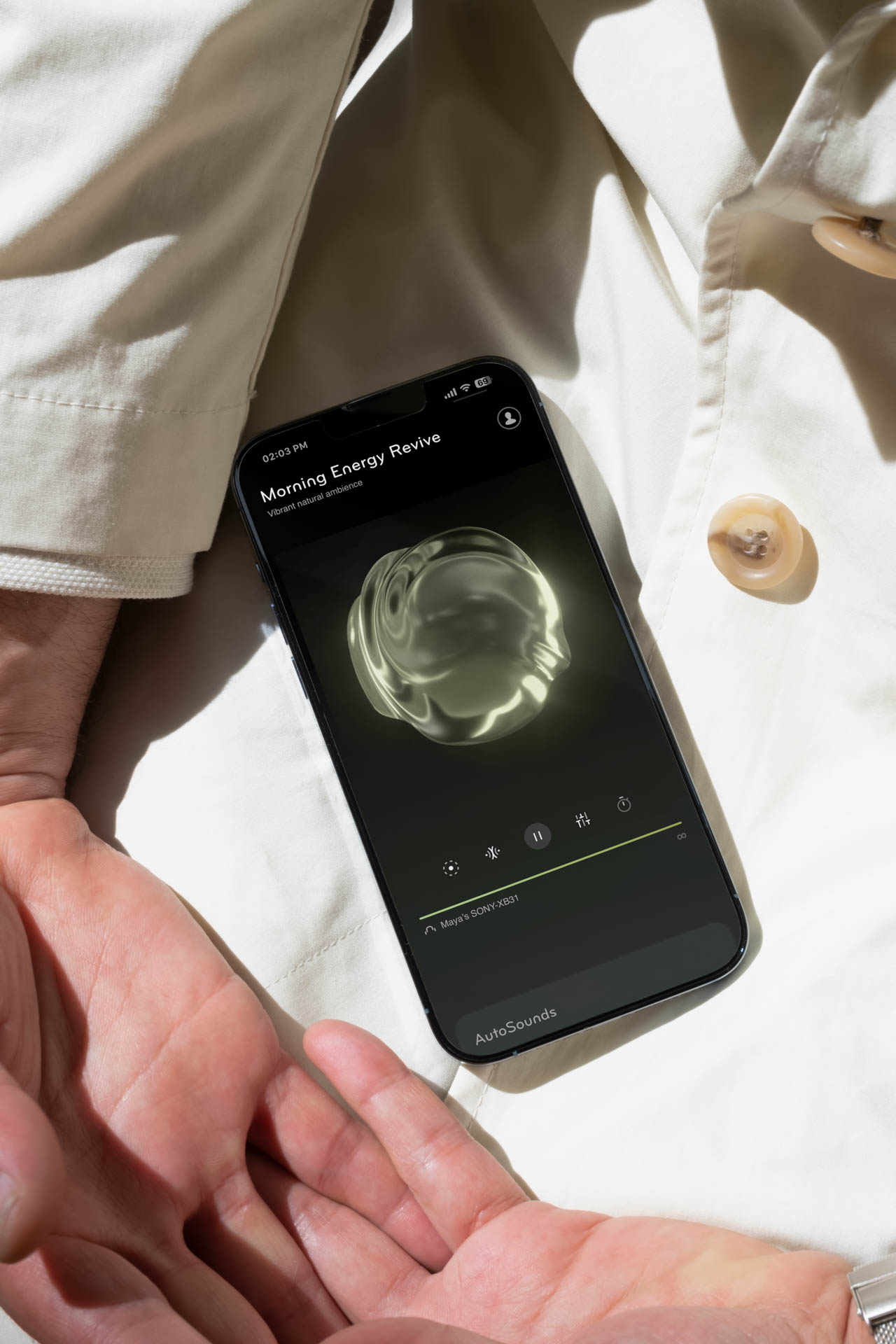 A black smartphone displays a music player interface titled “Modern Energy Revive” showing a circular, translucent graphic on its screen. The screen also indicates “Alive & Sync 5.0” and “AutoSounds” at the bottom. The phone rests on two hands, partially covered by light-colored fabric, with a portion of a person’s sleeve visible on the left. The background is blurred and out of focus.