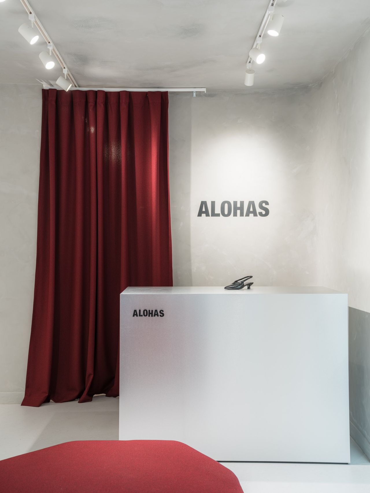 Minimalist ALOHAS retail store interior featuring a white display counter with logo signage, a single shoe on display, and a deep red curtain backdrop under track lighting.