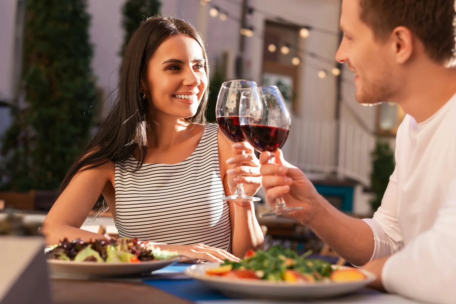Couple toasting with red wine glasses at an outdoor dinner table with fresh salads and plants in background.