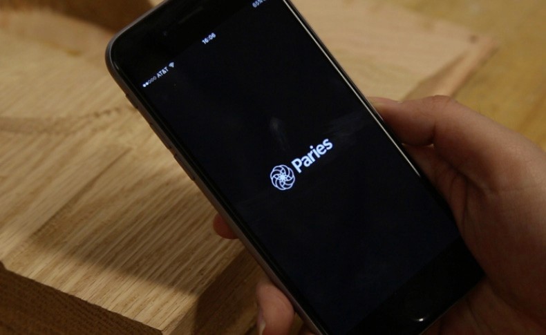 A hand holds a black smartphone displaying a social media app interface showing posts from two users, @banikunprakert and @beryoung, on a light brown wooden surface.
