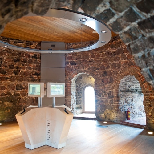 Interior of a stone-walled building with vaulted ceilings, featuring a modern information kiosk with two screens and a circular light fixture.