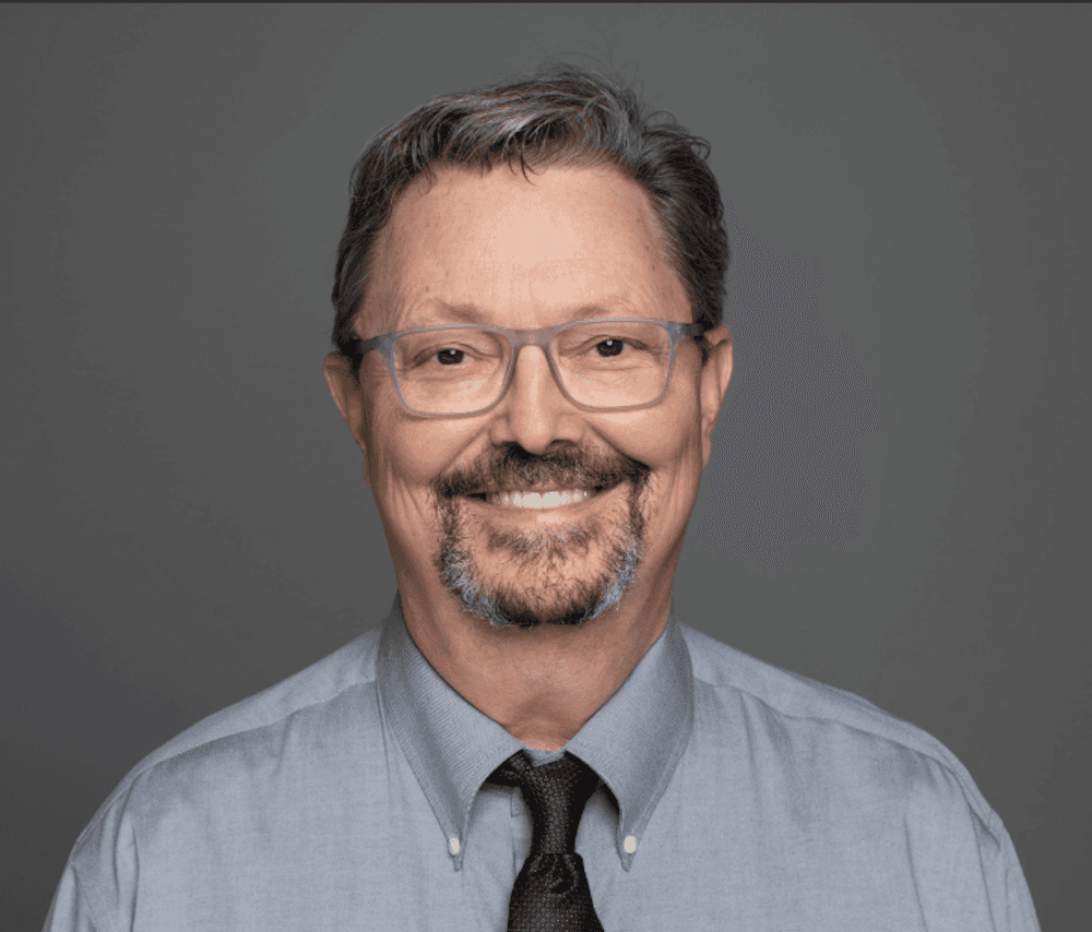 Dr. Michael Hutchison wearing glasses, a gray dress shirt, and black tie against a gray background.