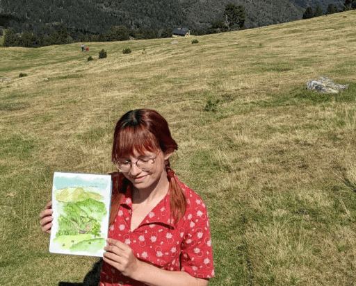 A person with braided red hair, wearing a red patterned shirt, stands on a grassy hillside holding a watercolor painting depicting a similar landscape, with distant mountains and trees in the background.