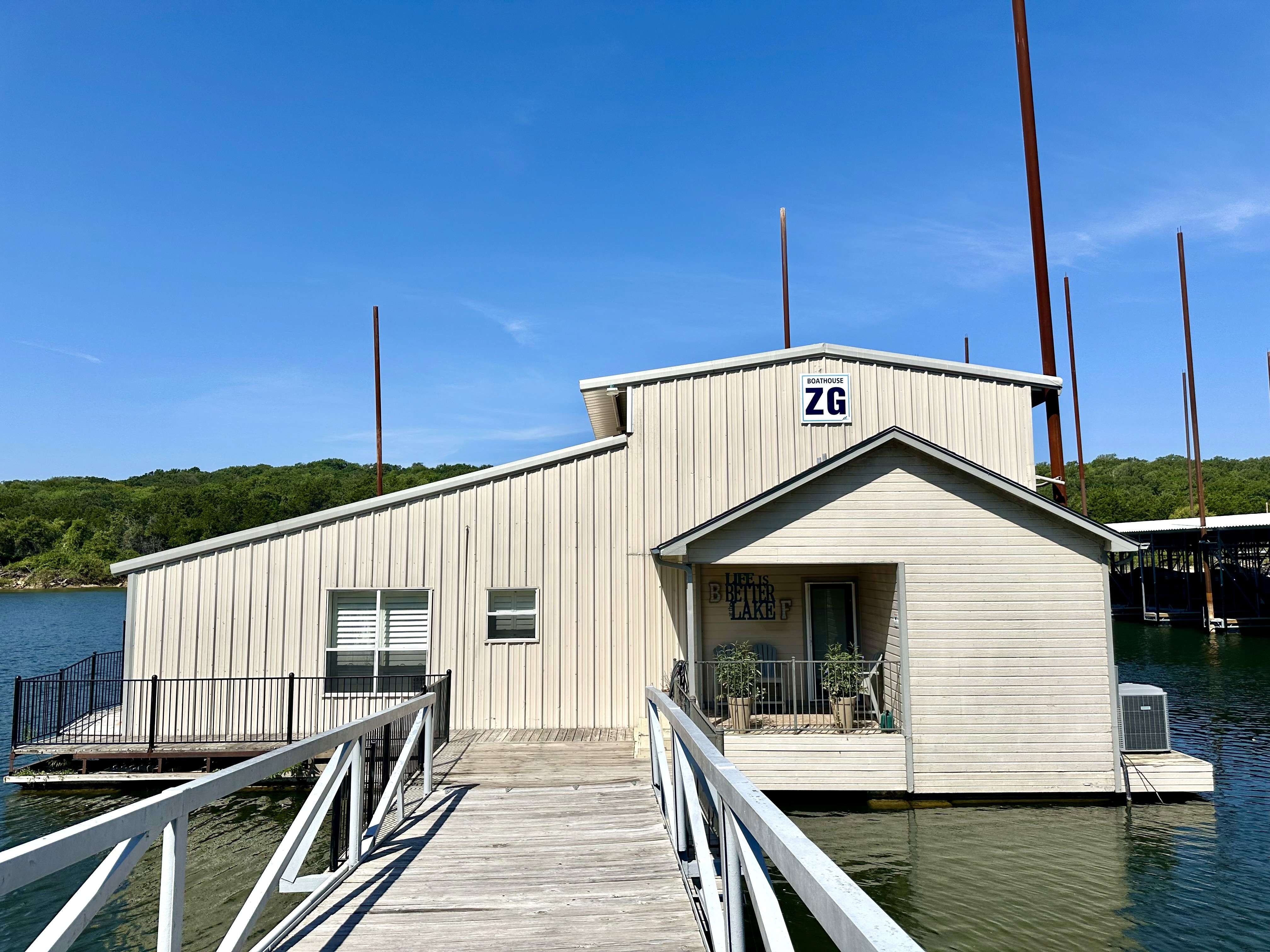 A beige boathouse labeled "ZG" sits on a clear lake, with a white wooden walkway leading to its entrance, against a backdrop of green trees and a bright blue sky.