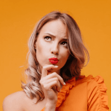 A thoughtful person with long blonde hair wearing a ruffled orange shirt against a vibrant orange background, rests their chin on their hand with a curious expression.