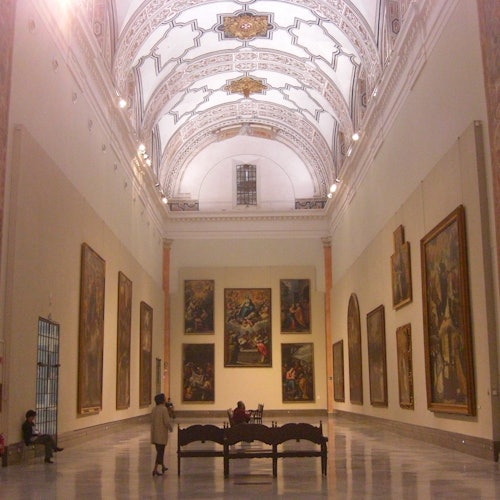 Une grande salle dans un musée d'art avec des arches ornées, de grands tableaux sur les murs et quelques personnes qui observent.