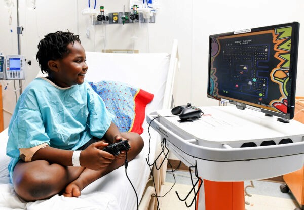 Kid sitting on a hospital bed, smiling and playing PacMan on a GO Kart.
