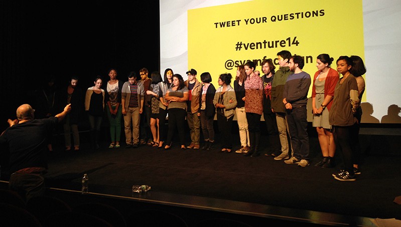 A photo of a group of people sitting on a stage and listening to a lecture, while behind them on a projector screen a yellow background with black text is set.