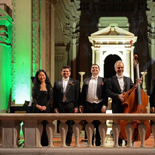 Five musicians in formal attire stand on a stage with ornate architectural décor, including green lighting and a white arch in the background.
