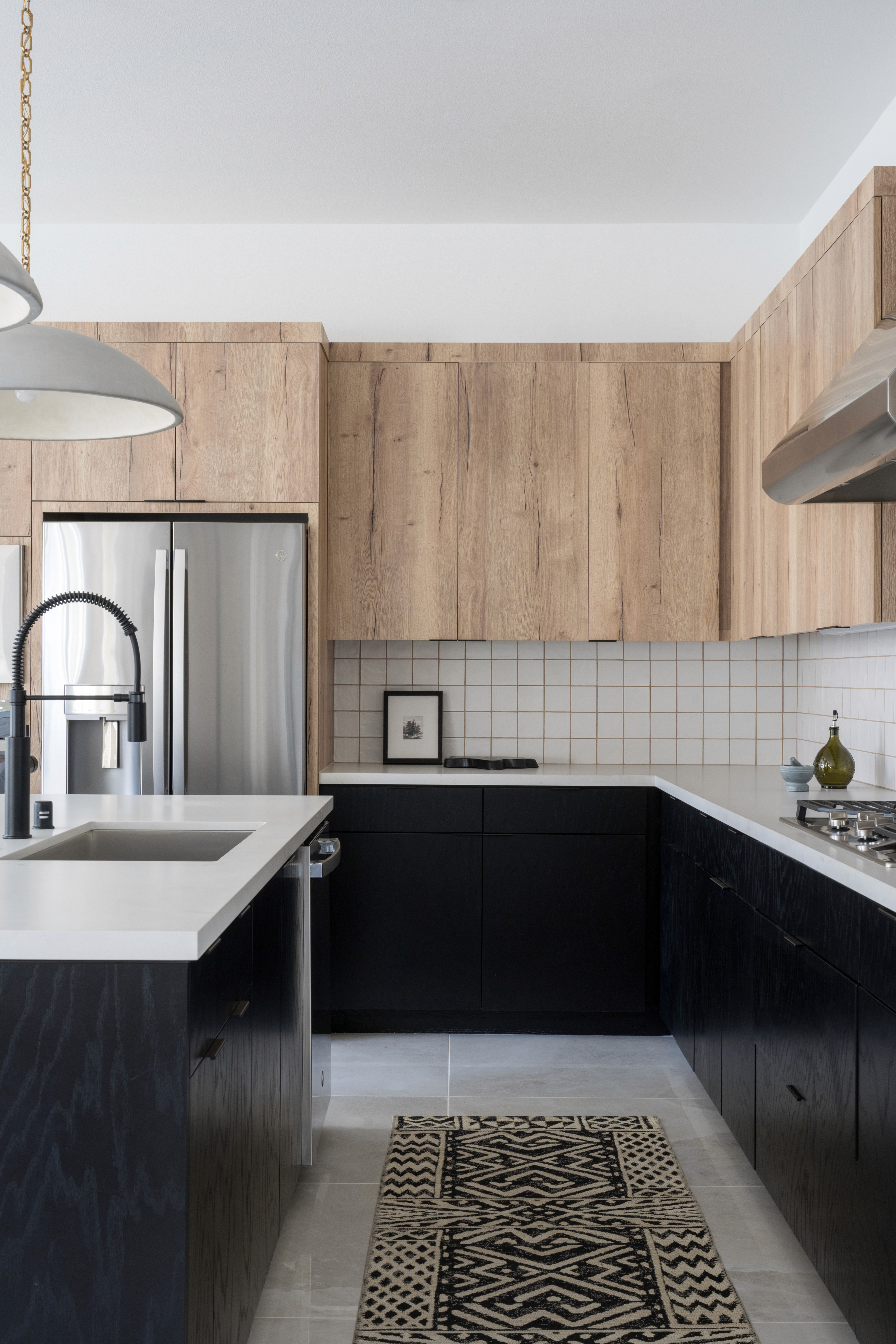 Moody kitchen with high, neutral wood cabinets and low, black cabinets, white countertops, stainless steel appliances, grey tiled floors, and black and white patterned runner rug  by Jubilee Interiors in Arcadia, California
