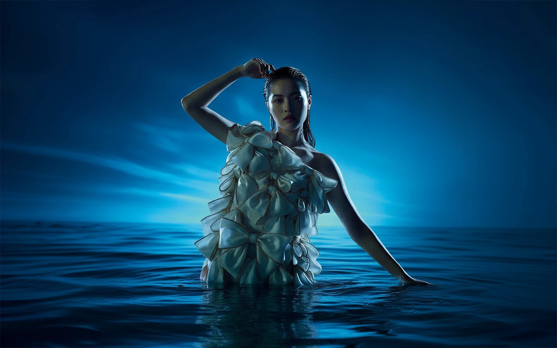 A woman in a sculptural dress stands in water under dramatic blue lighting, creating a surreal and artistic visual effect.