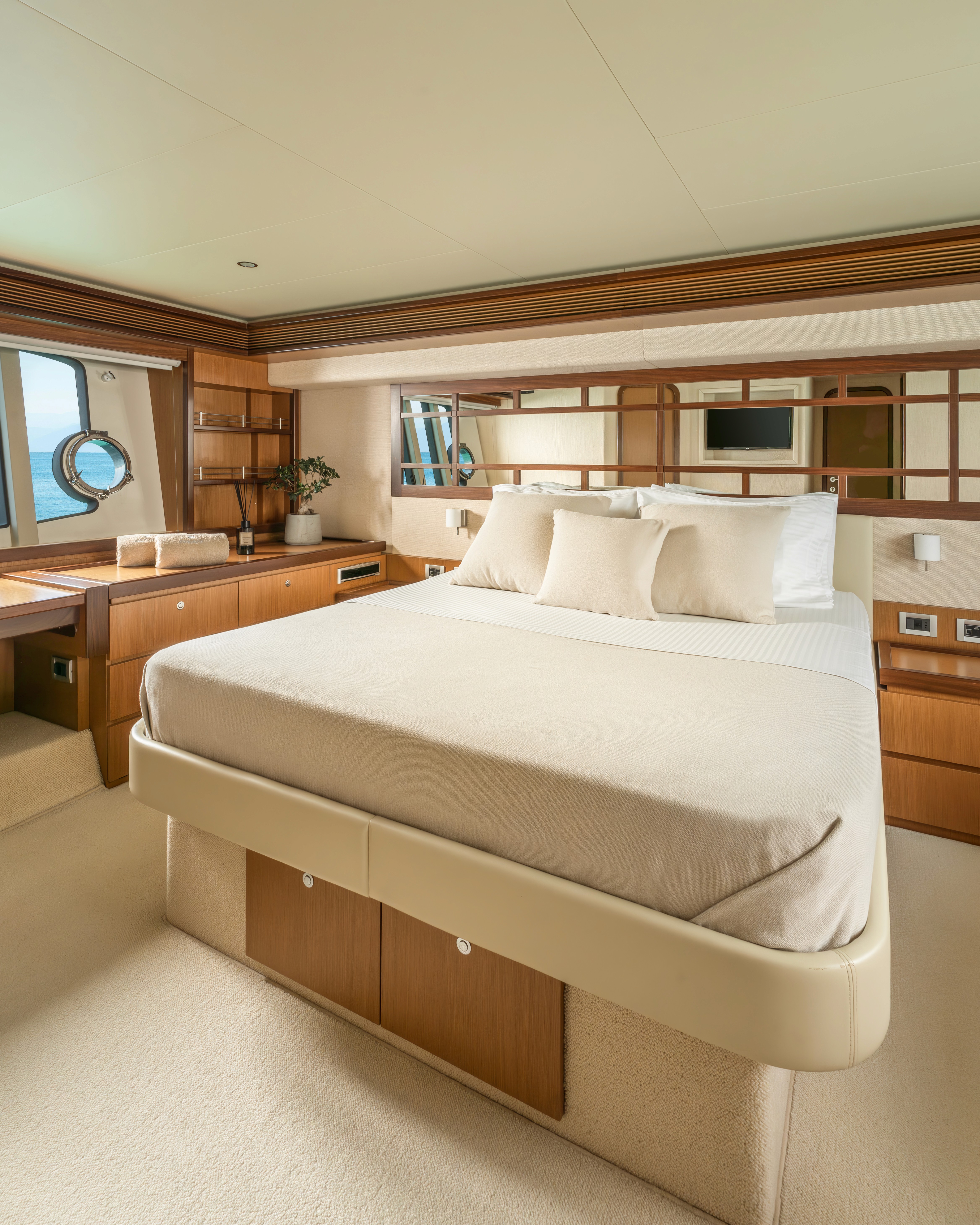 Luxurious yacht master cabin featuring a queen bed with cream linens, warm teak wood cabinetry, and ocean view porthole with nautical decor.