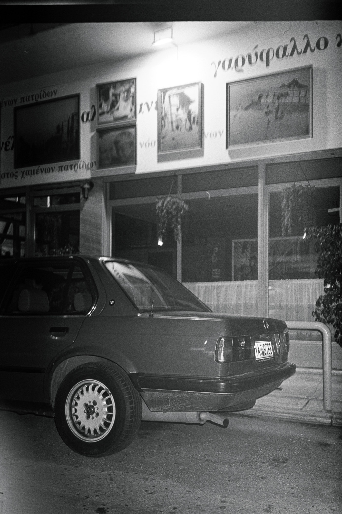 A black and white photo of a car parked outside a building with framed pictures and signs written in Greek above large windows.