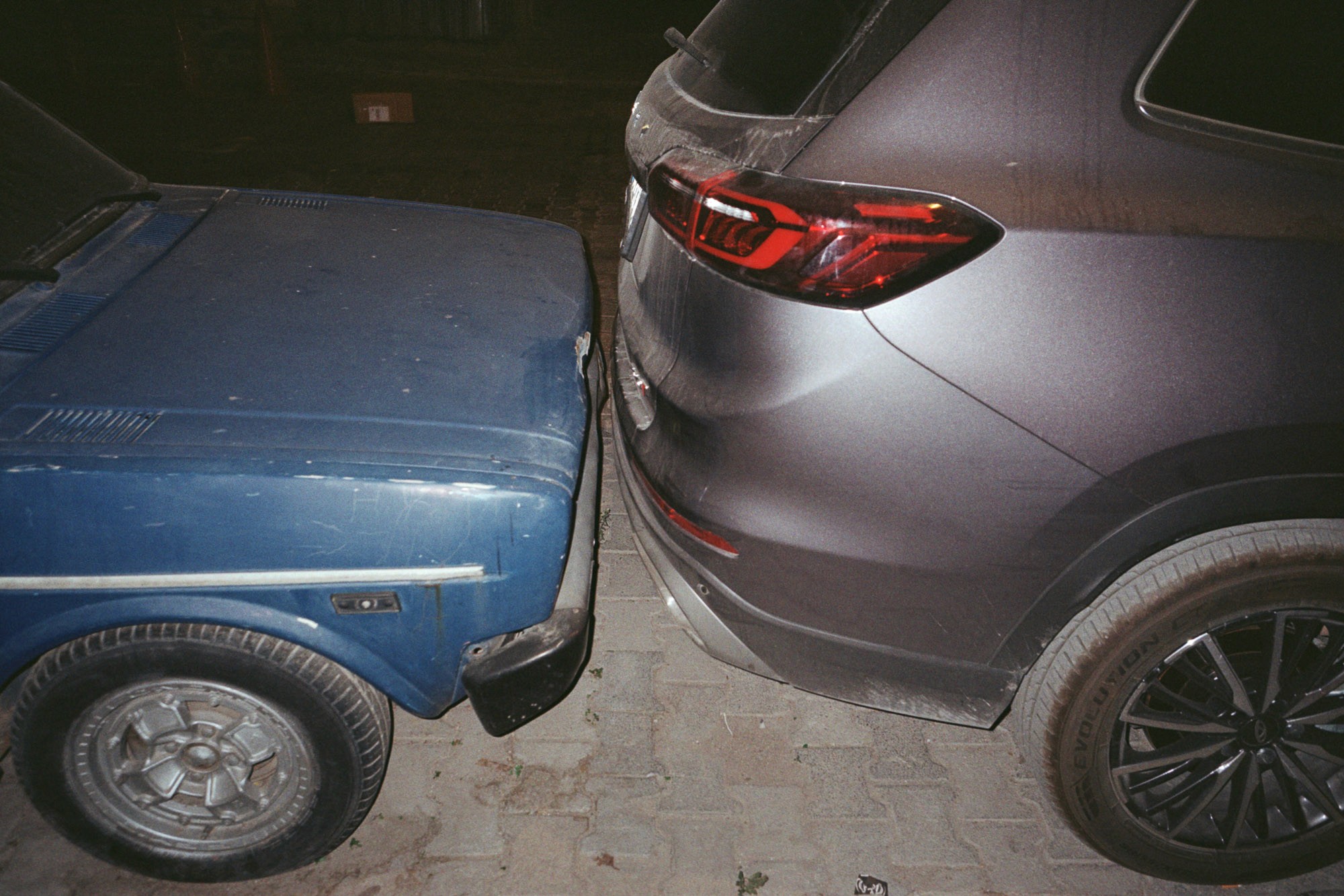 A close-up image shows an old blue classic car parked very closely to a modern dark gray SUV, almost touching, on a paved surface at night.