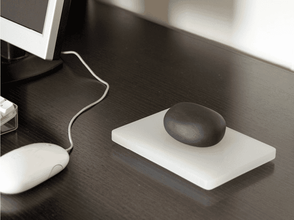 A dark wooden desk holds a white computer mouse connected by a white cord to a computer monitor. A dark stone rests on a white rectangular pad placed on the desk. The scene is captured in black and white.