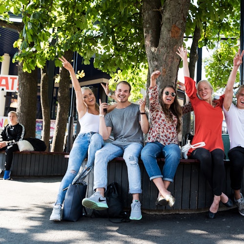 Five joyful people sitting on a bench under trees, raising their arms and smiling. Another person sits on a nearby bench.