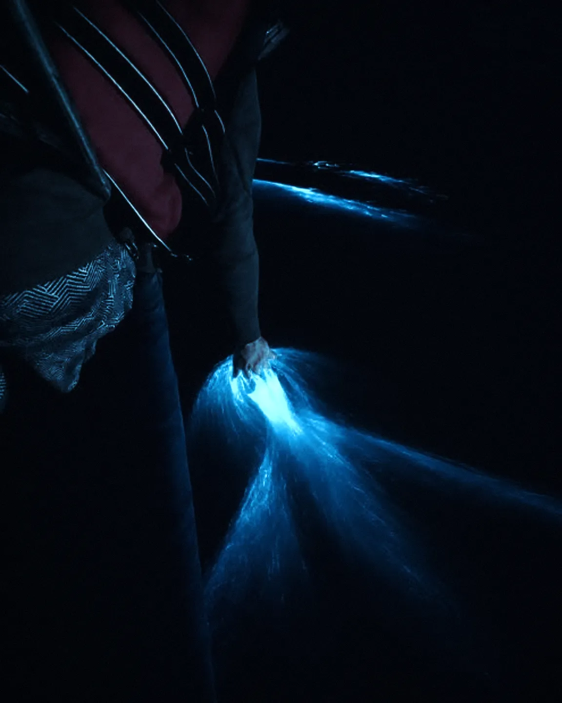 Hand moving through dark water that glows blue with bioluminescence at night
