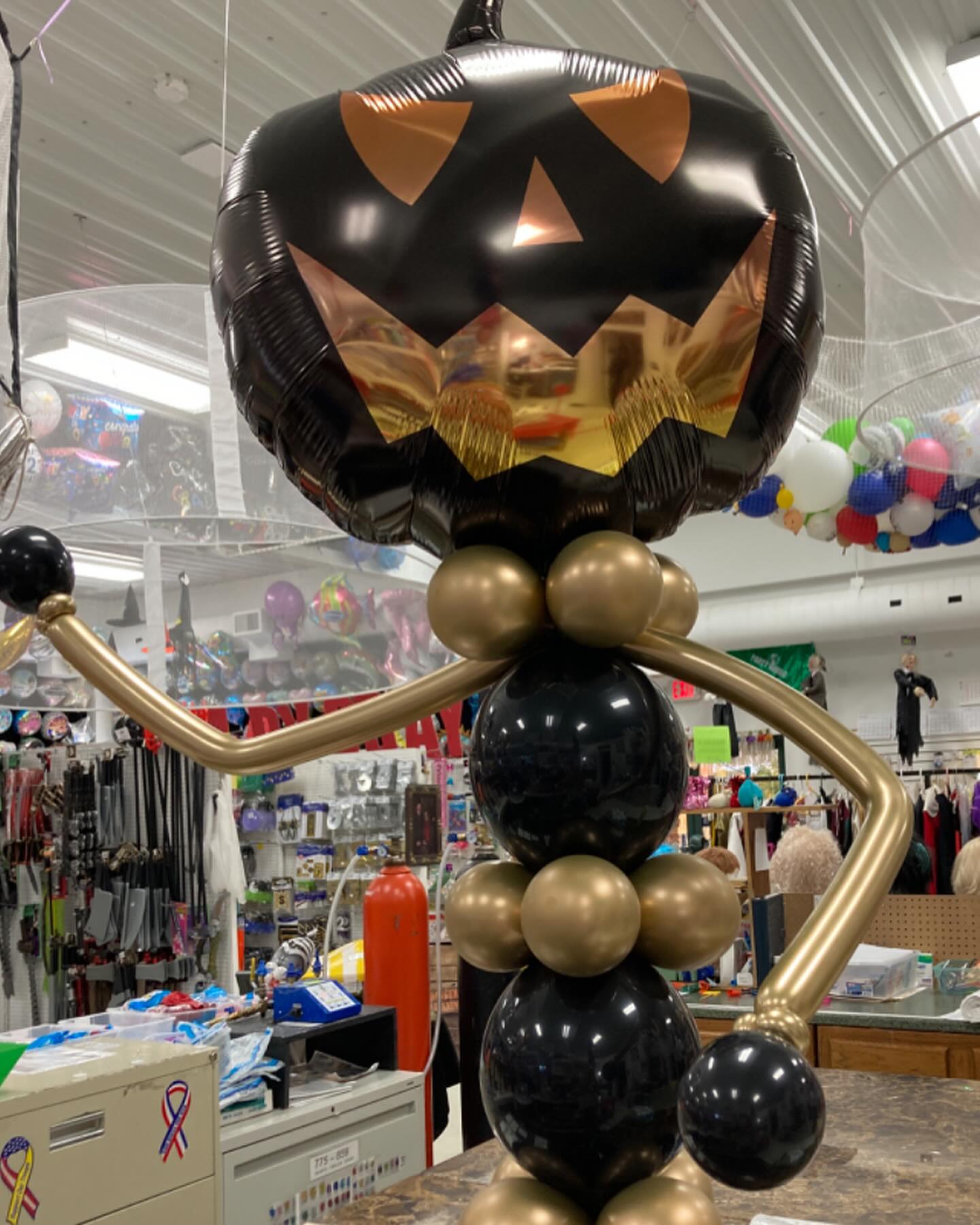 A festive Halloween balloon sculpture featuring a metallic black and gold jack-o'-lantern head on a tall body made of black and gold balloons, displayed in a colorful store filled with various party supplies and decorations.