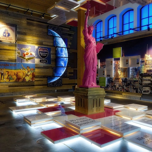 Indoor exhibit featuring a pink Statue of Liberty replica surrounded by illuminated display panels and various wall posters.