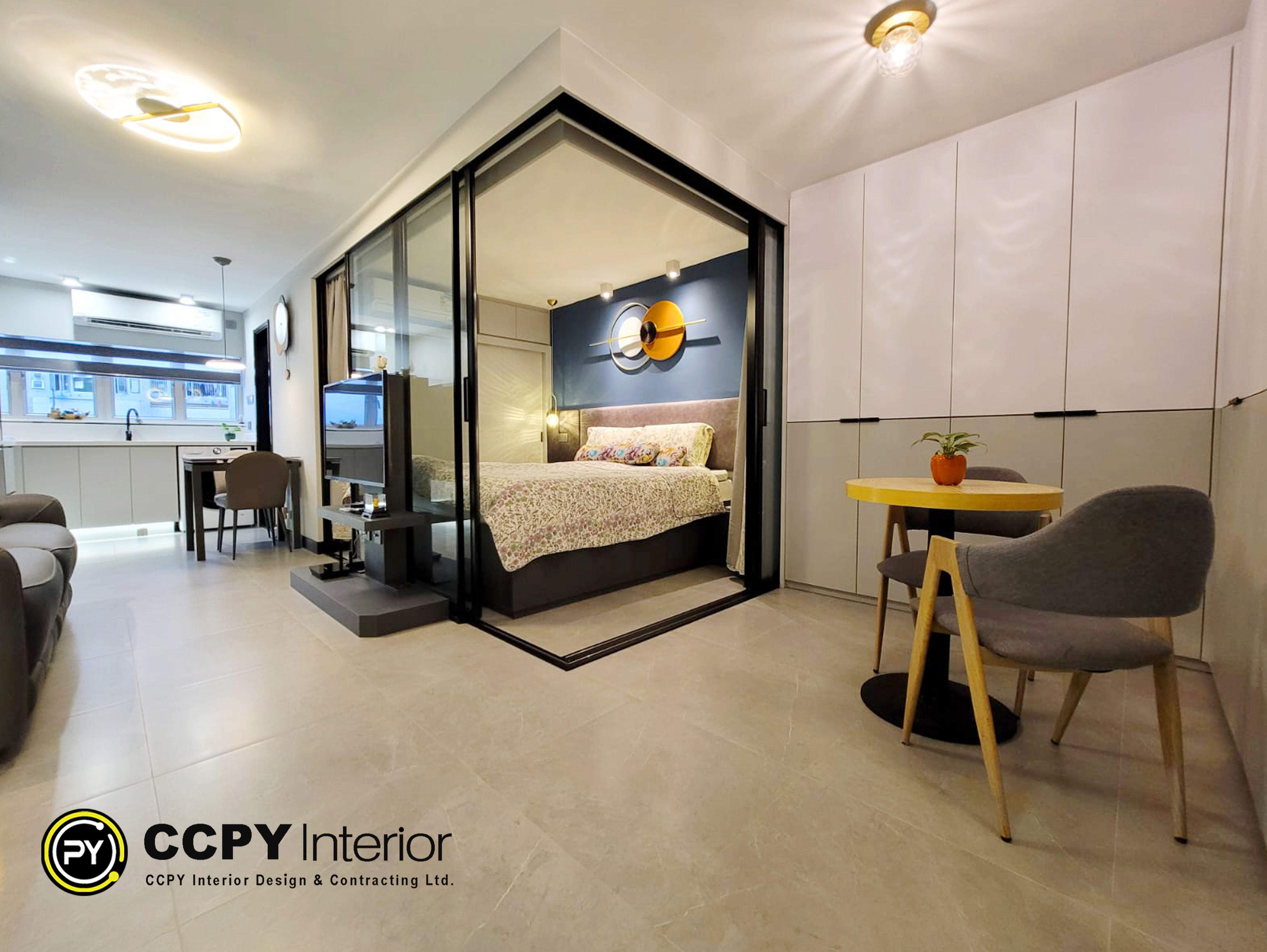 This modern contemporary residence features a cleverly partitioned bedroom, utilizing a sleek, dark-framed glass sliding door to promote an airy, light-filled atmosphere. The thoughtfully designed floor plan includes a custom built-in entertainment console and a cozy dining nook anchored by a circular table, all tied together by a sophisticated neutral palette and warm, integrated lighting.