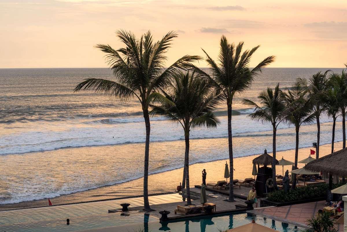 Palm trees line a beach and pool area at sunset with waves breaking on the shore under an orange sky.