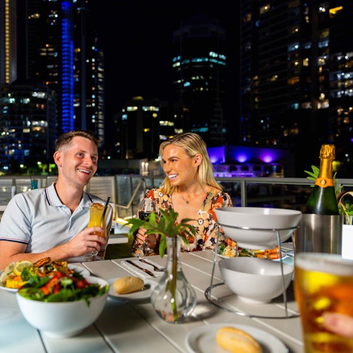 A couple enjoying dinner at an outdoor restaurant at night, surrounded by city lights and skyscrapers, with drinks and food on the table.