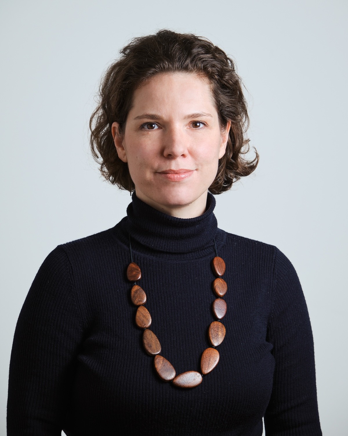 Eine Frau mit lockigem Haar, die einen schwarzen Rollkragenpullover und eine Perlenkette trägt, steht vor einem hellen Hintergrund.