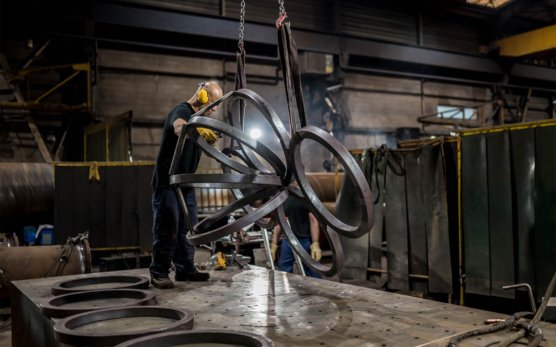 Dans un atelier industriel sombre, un ouvrier portant un casque de protection et des gants travaille sur une structure métallique complexe composée de plusieurs anneaux soudés, suspendue par des sangles. L'homme est concentré sur son travail, tandis que d'autres anneaux métalliques sont disposés à plat sur une grande table de travail robuste au premier plan. L'environnement est celui d'une usine de métallurgie, avec des écrans de protection et des outils de soudure visibles en arrière-plan.