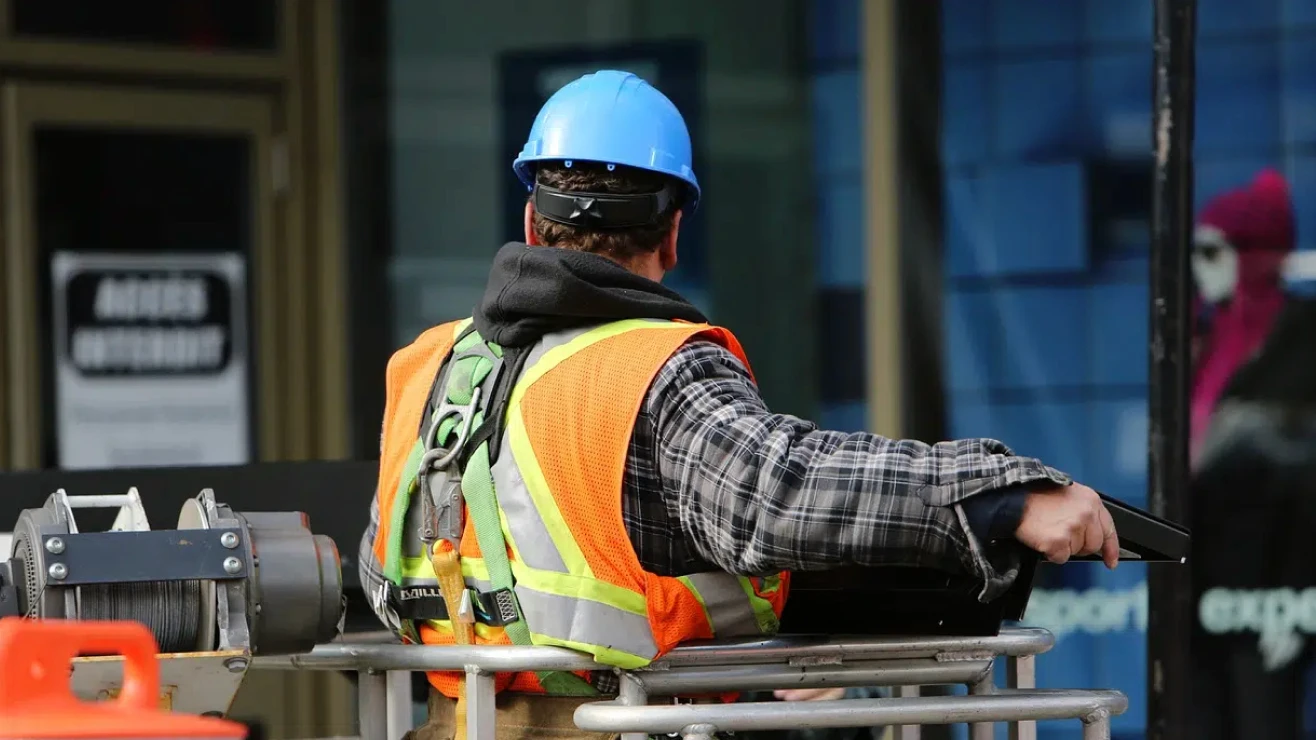 Pessoa usando capacete azul, colete refletivo e cinto de segurança em uma plataforma de trabalho elevada, diante da fachada de um prédio.