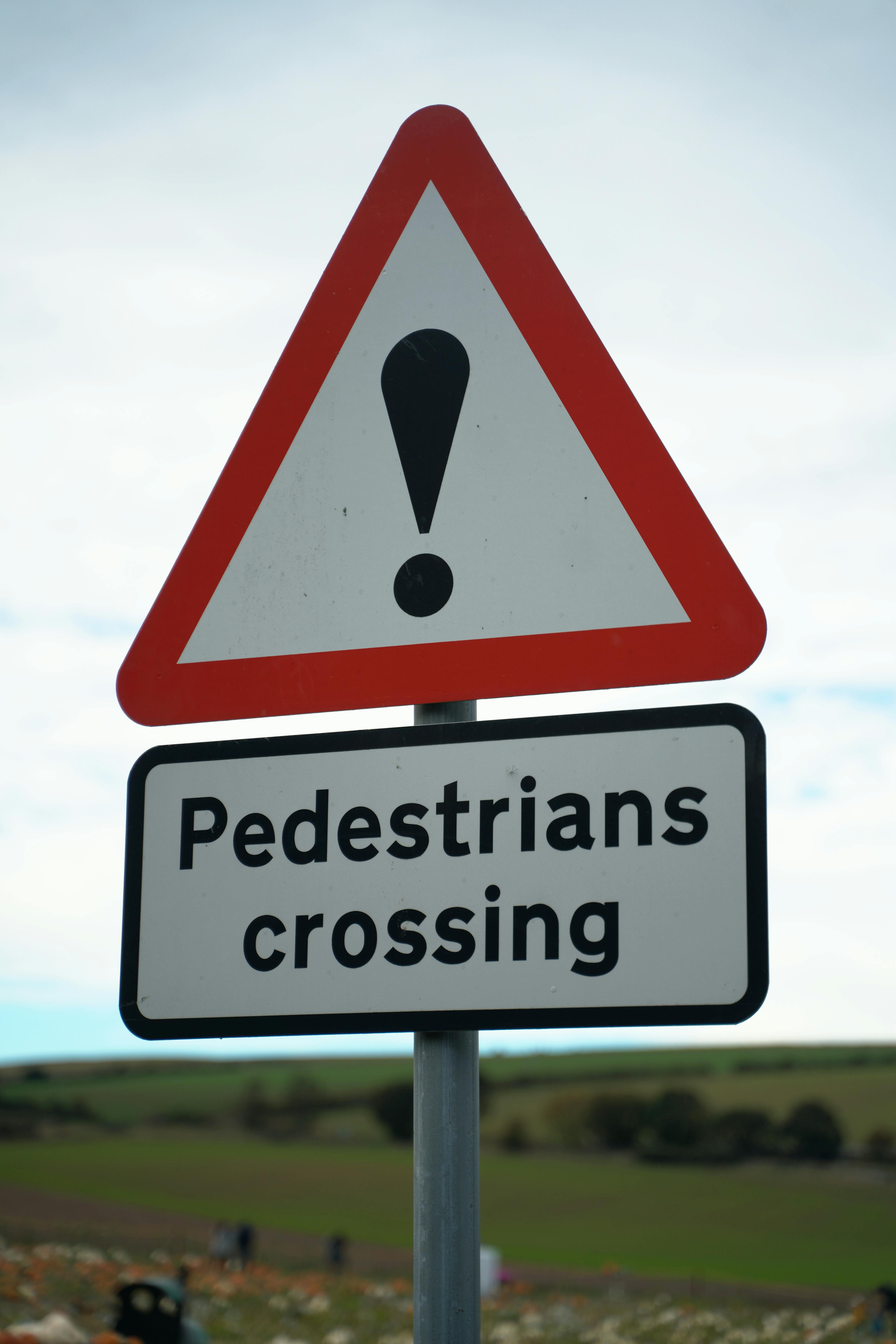 UK road sign showing a red warning triangle with an exclamation mark and a plate reading “Pedestrians crossing.”