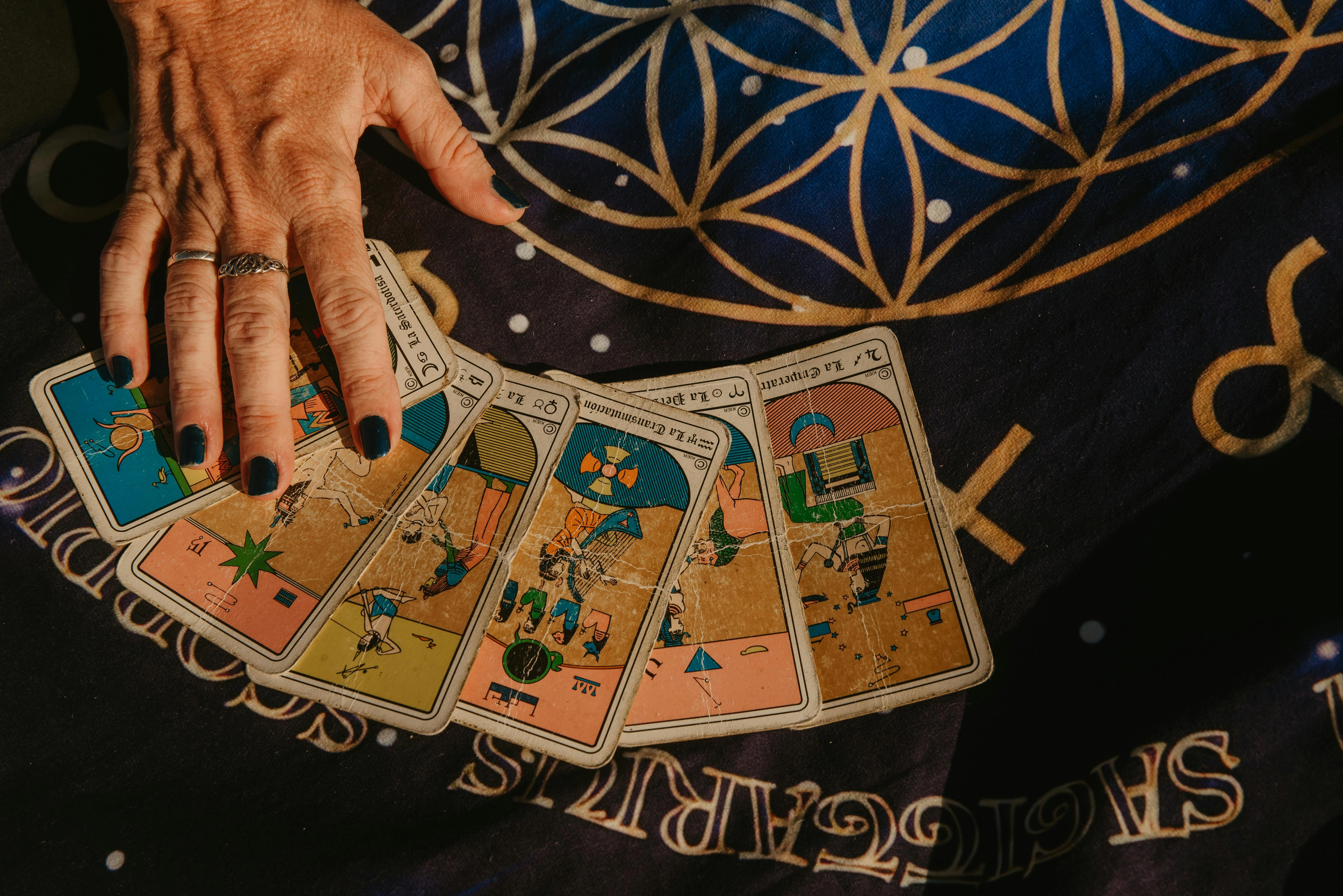 A close-up of a hand with silver rings fanning out vintage tarot cards on a blue astrological cloth with gold sacred geometry symbols.