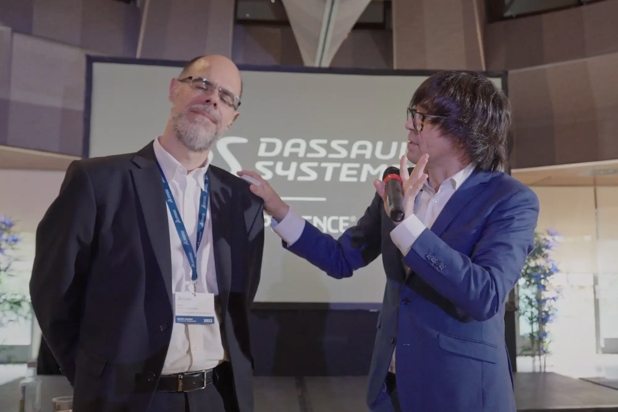 Portfolio Thankium: Dos hombres en un evento de Dassault Systèmes, uno de ellos sosteniendo micrófono, pantalla de fondo.