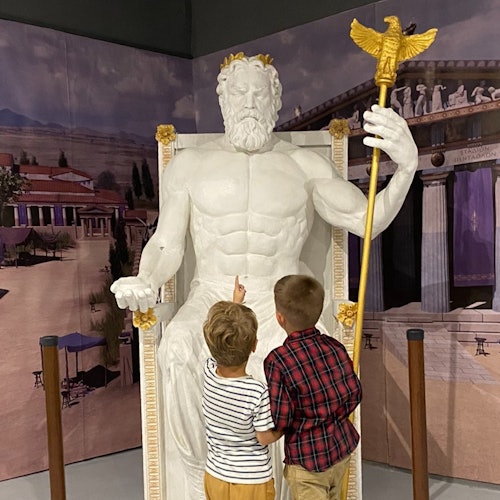 兩名小朋友站在一座巨大的坐姿鬍鬚人物雕像前，指着它。背景是一幅古老建築的壁畫。
