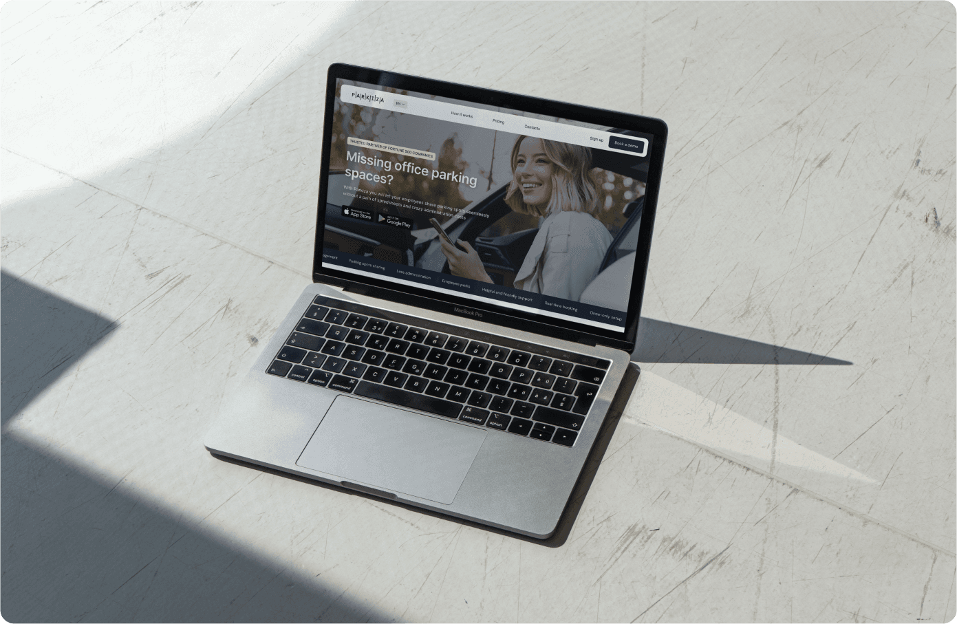 A MacBook Pro laptop on a white marble surface displaying the Parkiza website. The website shows a banner with a smiling woman using her phone and text that reads: 'Missing office parking spaces? Parkiza helps you and your employees share, track and manage parking spaces seamlessly from your phone or desktop.' There are buttons for downloading the app from the App Store and Google Play. The top navigation bar includes options for 'How it works,' 'Pricing,' 'Resources,' 'About,' 'Contacts,' and a 'Sign up' button. There is also a 'Book a demo' button on the right side of the banner.