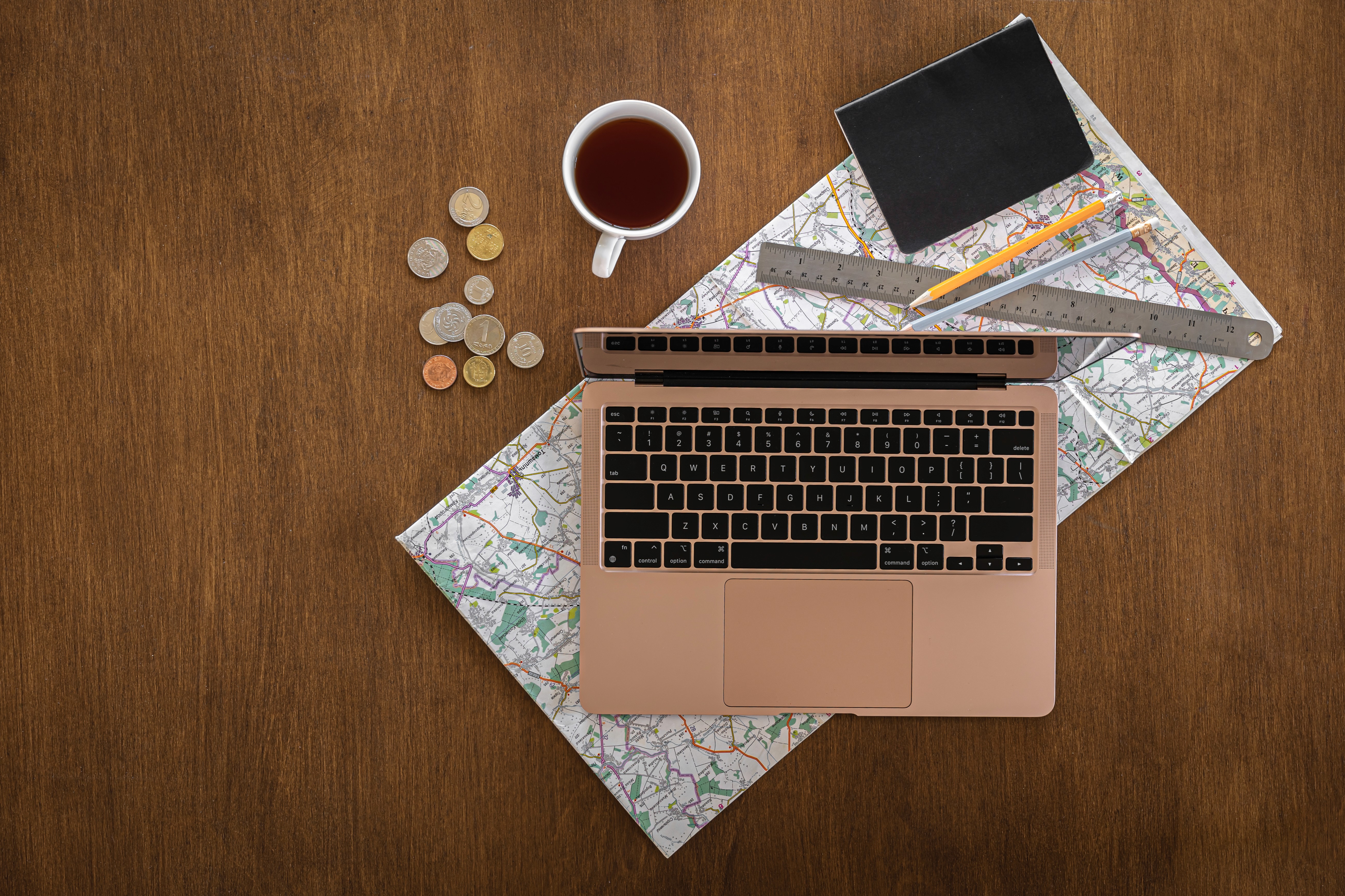 Flat lay of travel planning essentials with a laptop, paper map, coins, notebook, rulers, and a cup of tea on a wooden desk.