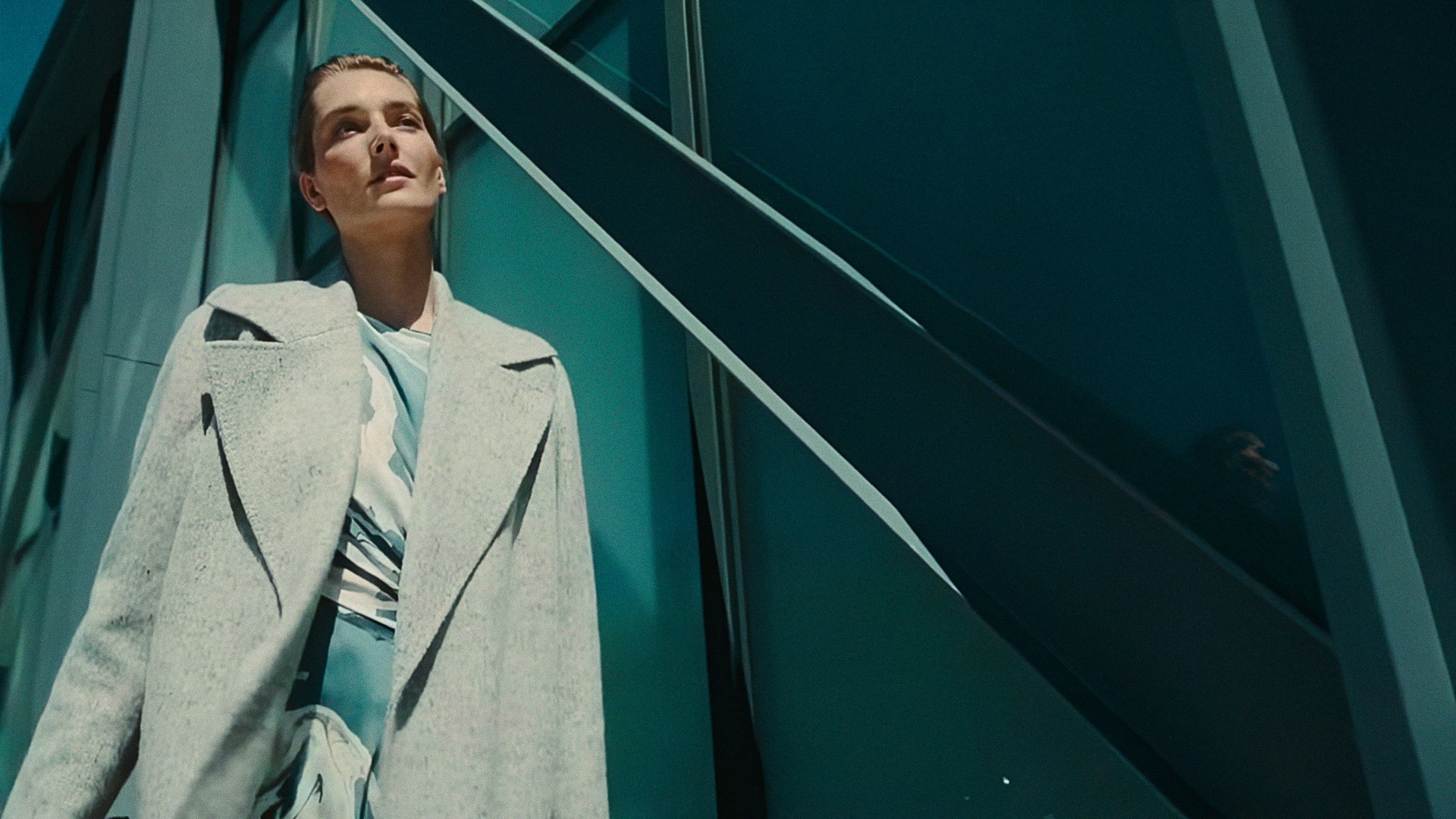 A woman in a gray coat among teal steel beams, looking up, Lafayette 148 New York