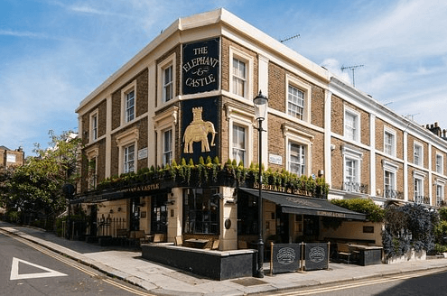 A smiling man with curly hair is sitting at a desk, looking at the camera, with a laptop and a lamp in the background.