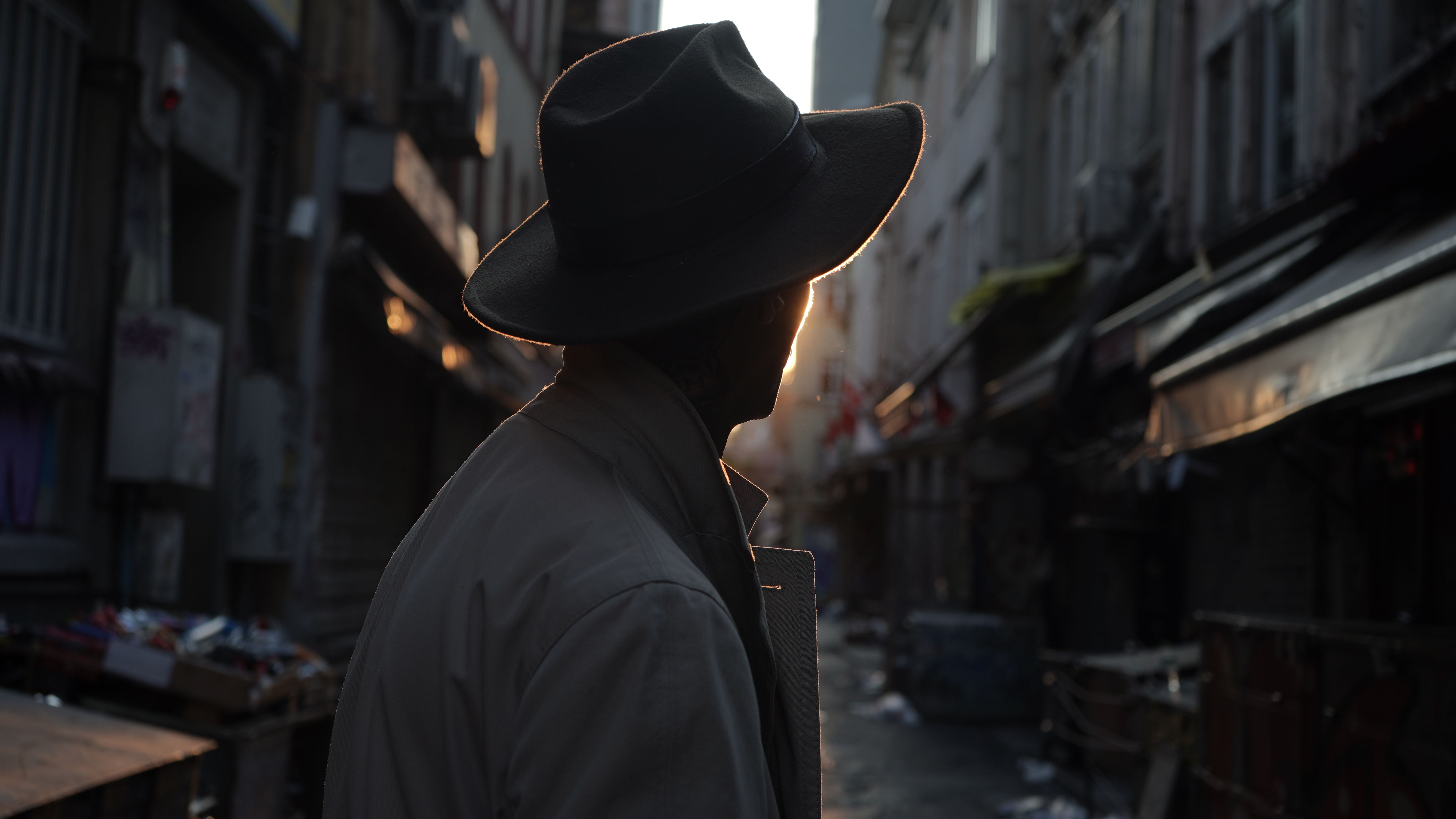 A silhouette of a man wearing a fedora in a backlit alley at sunrise.