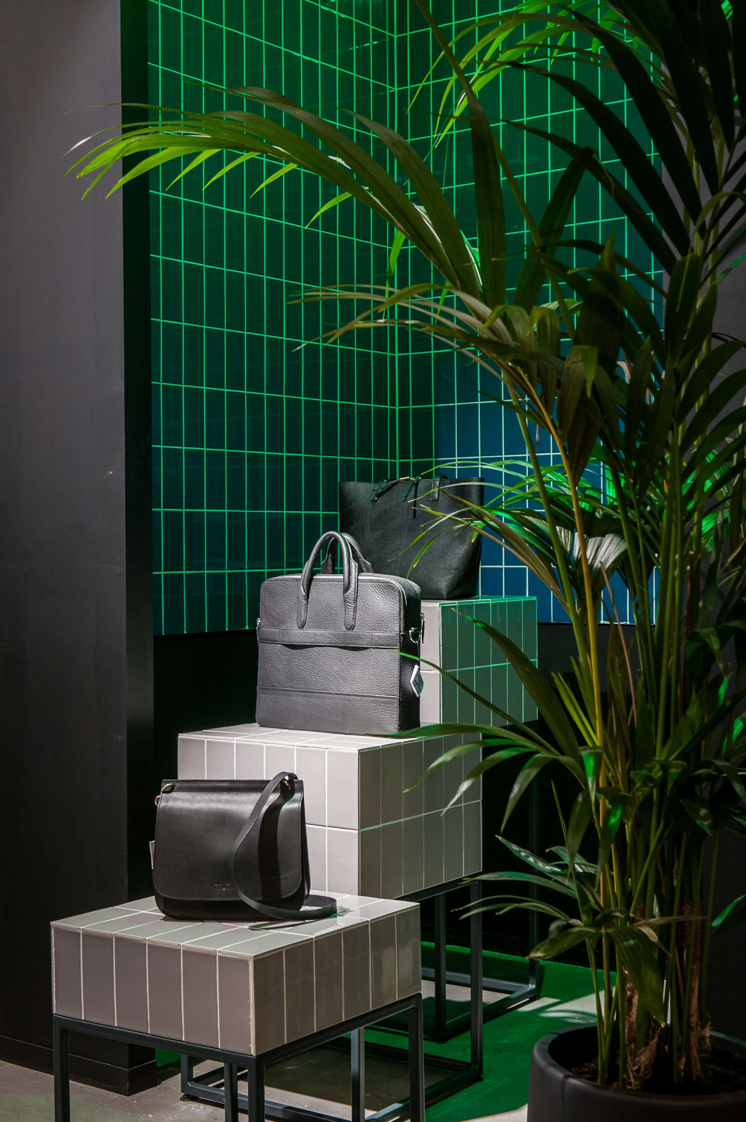 Leather bags displayed on white tiled pedestals in front of a glowing green grid wall at the 17 FASHION BRANDS showroom, framed by a large palm plant.