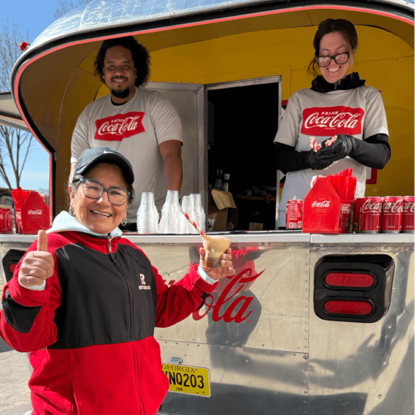 A smiling woman holds a drink and gives a thumbs-up in front of a retro Coca-Cola truck, where two workers in Coca-Cola shirts stand ready to serve drinks and snacks.