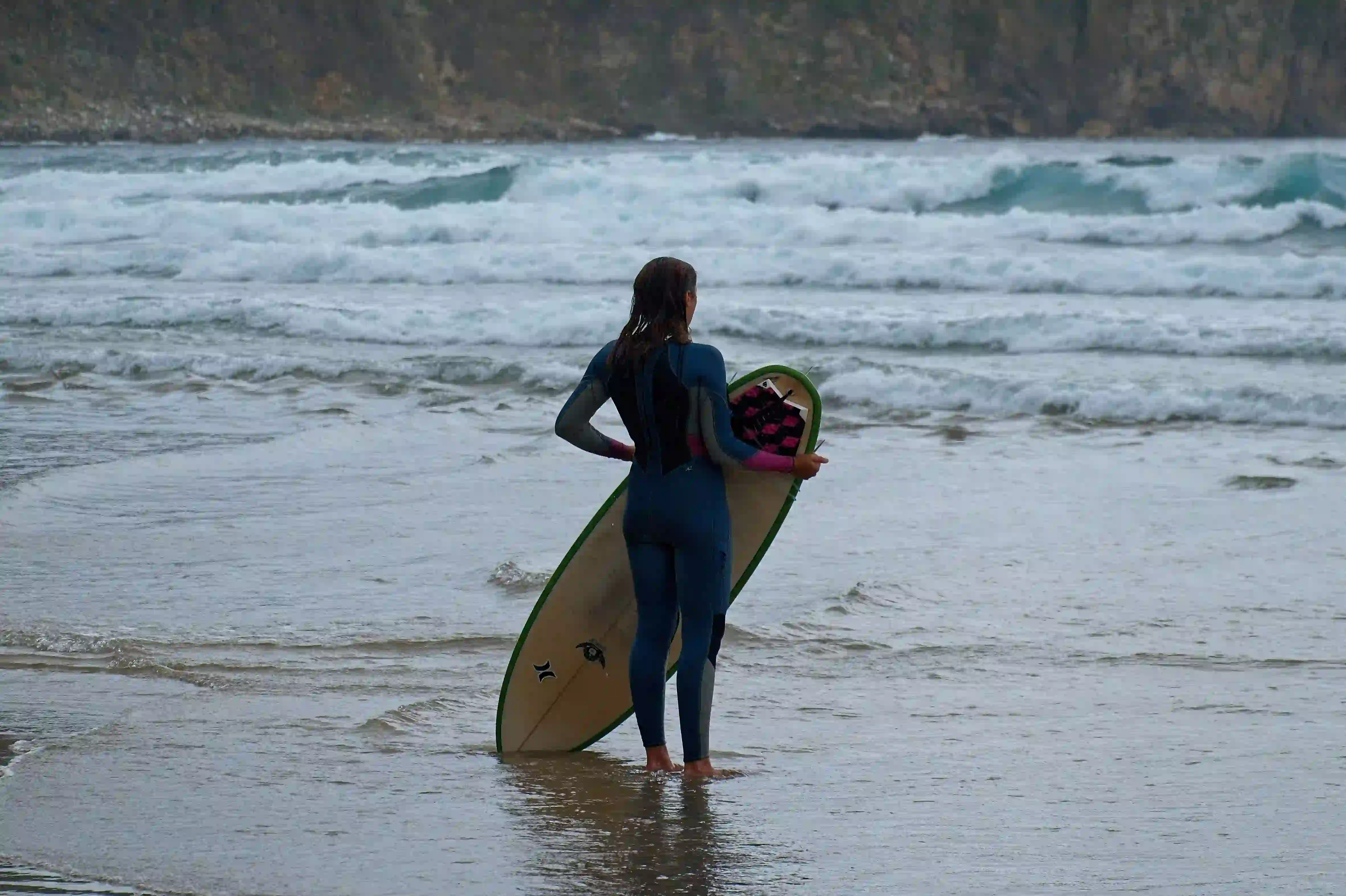 Man in a wetsuit standing in shallow water holding a surfboard and watching incoming waves.