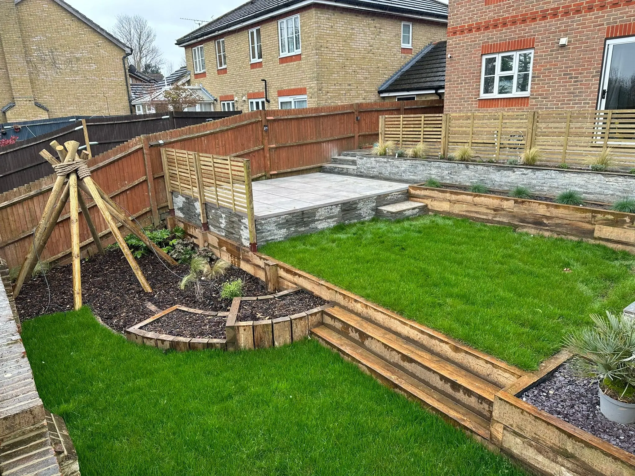 A well-maintained garden featuring green grass, wooden pathways, and a wooden structure against a backdrop of houses.